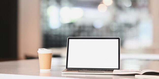 Computador portátil, café, caderno e caneta na mesa de escritório branco.