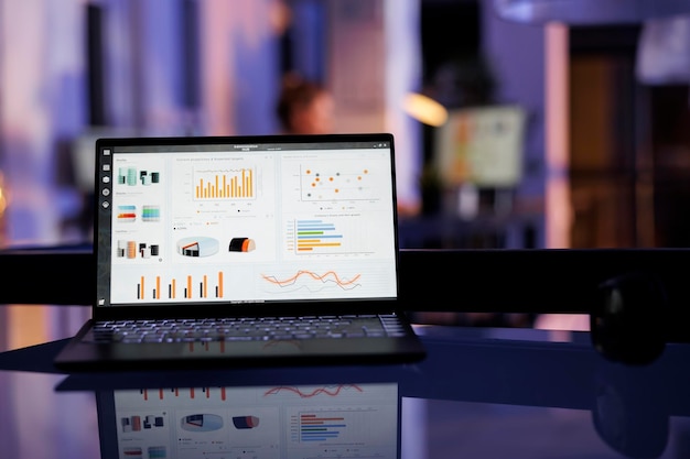 Computador portátil com estatísticas de marketing de pé na mesa de escritório no escritório de inicialização, em fundo empresário trabalhando horas extras no relatório de crescimento financeiro, digitando a estratégia da empresa. Conceito de negócio