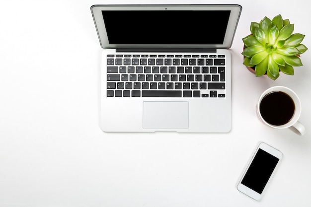 Computador portátil com planta em uma panela, telefone móvel e xícara de café na superfície branca.