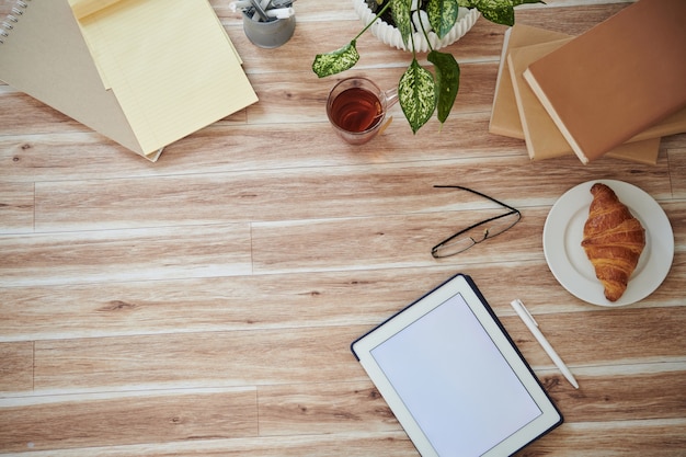 Computador tablet com tela vazia, livros, croissant e copos na mesa, vista de cima