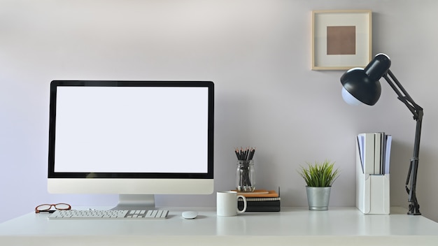 Computadora de pantalla vacía de espacio de trabajo moderno y accesorios creativos con taza de café y lámpara en mesa blanca.