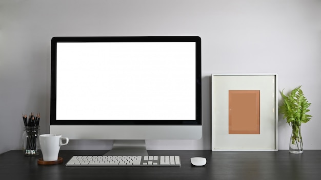 Computadora personal del espacio de trabajo con pantalla en blanco, portalápices, marco, planta en maceta y taza de café que se juntan en una mesa de madera moderna con pared blanca