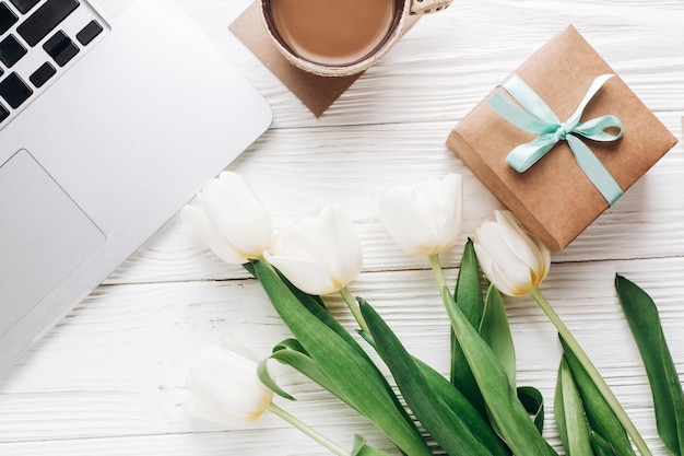 Computadora portátil con café de la mañana y tulipanes y elegante caja de regalo sobre fondo rústico de madera blanca, dispositivos de trabajo independientes planos con espacio para texto, vacaciones de primavera, feliz día de la mujer