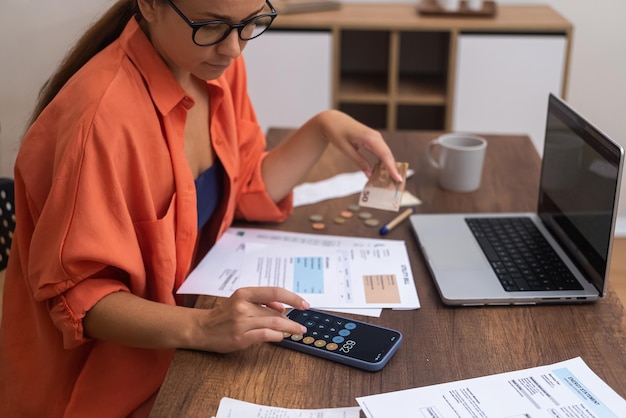 Con una computadora portátil cerca, una mujer calcula eficientemente las finanzas del hogar en su teléfono inteligente en un escritorio