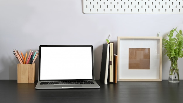 Computadora portátil en el escritorio de trabajo junto con la computadora de pantalla en blanco blanca, libros, cuaderno, portalápices, marco de fotos, planta en maceta que se junta con pared de cemento blanco.