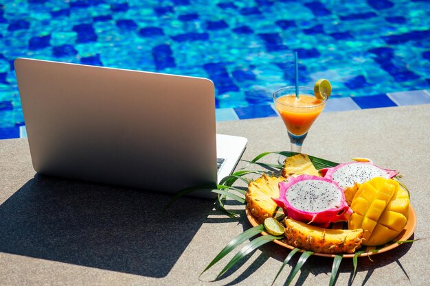 Computadora portátil con golpes de frutas tropicales frescas papaya, piña, fruta del dragón por piscina trabajo a distancia freelance y frilansing