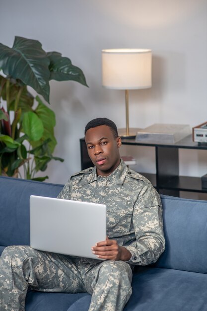 Computadora portátil, info. Atractivo joven militar afroamericano serio con portátil sentado en el sofá en casa