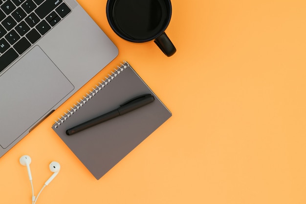 Computadora portátil moderna, auriculares blancos, cuaderno gris con un bolígrafo y una taza de café en la superficie naranja