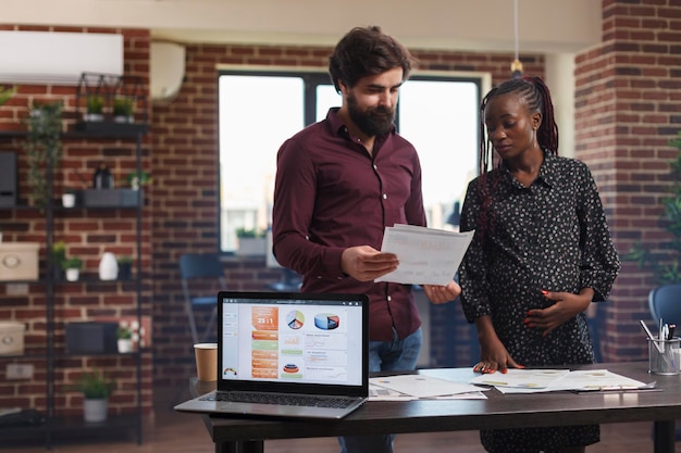 Computadora portátil moderna en el lugar de trabajo con gráficos de análisis de negocios en pantalla. Empresaria embarazada afroamericana y compañera de trabajo analizando los ingresos anuales de la empresa y el papeleo contable.