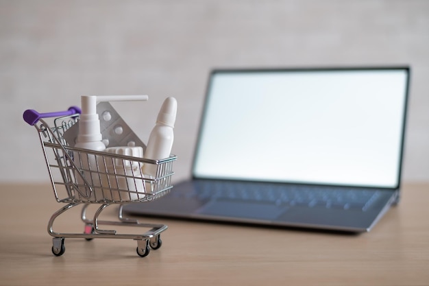 Computadora portátil con pantalla blanca en blanco y carrito de compras en miniatura lleno de medicamentos Concepto de farmacia en línea Sitio web para comprar tabletas