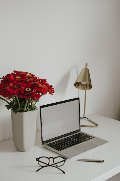 Computadora portátil con pantalla en blanco en la mesa con lámpara de gafas ramo de flores de gerber rojo Plantilla de diseño de interiores de espacio de trabajo de estilo influenciador estético con espacio de copia de maqueta