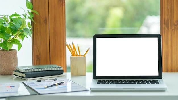 Foto computadora portátil con papelería de pantalla vacía y planta en maceta sobre una mesa blanca en una oficina luminosa