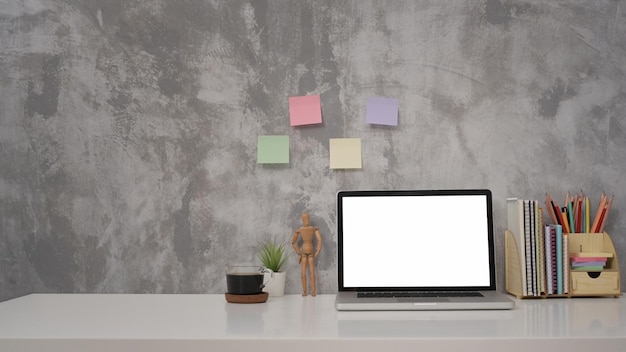 Computadora portátil de vista frontal con taza de café con pantalla en blanco y suministros en una mesa blanca contra un muro de hormigón