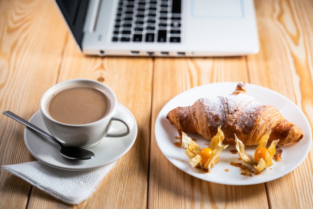 Computadora, taza de café y croissant en mesa de madera marrón