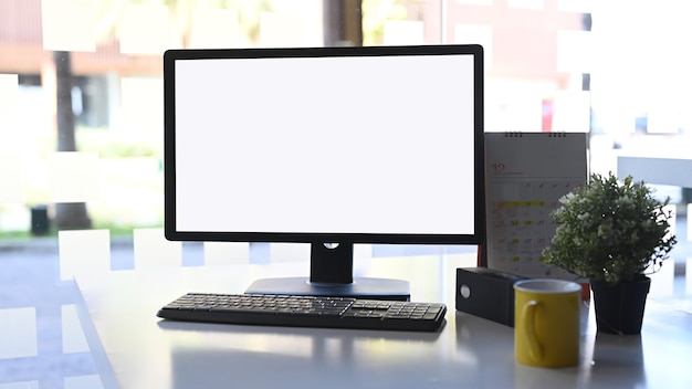 Computadora de vista frontal con pantalla en blanco, taza de café, planta y calendario en la mesa blanca.