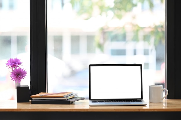 Computer-Laptop mit weißem leerem Bildschirm zusammen mit Kaffeetasse, Stapel Notizbuch und Topfpflanze auf dem modernen Holztisch mit beschnittenen schwarzen Fenstern