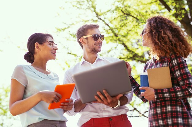 Comunicação agradável. Jovem exuberante segurando seu laptop e discutindo um novo projeto com seus colegas de grupo