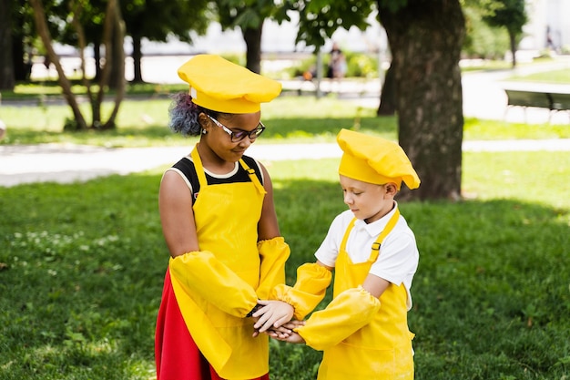 Comunicación entre amigos multirraciales de niños cocineros negros africanos y caucásicos con sombrero de chef amarillo y delantal tomándose de la mano y divirtiéndose juntos