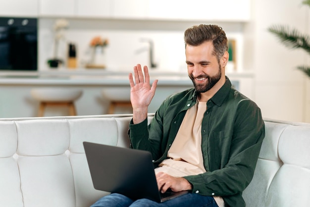 Comunicación distante Un hombre caucásico, guapo y elegante, se sienta en un sofá en una acogedora sala de estar, usa su computadora portátil hablando en línea por videoconferencia con amigos, hace que el gesto de saludo sonría