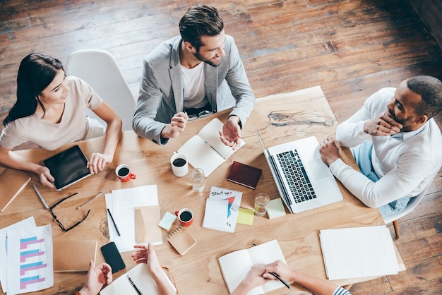 Comunicación de equipo. Parte de la vista superior del grupo de cinco personas discutiendo algo con una sonrisa mientras está sentado en la mesa de la oficina