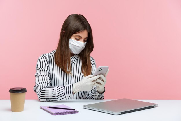 Comunicación en línea en cuarentena de coronavirus de autoaislamiento Mujer sentada segura y saludable con mascarilla facial higiénica y guantes protectores en el lugar de trabajo escribiendo mensajes usando una foto de estudio de teléfono móvil