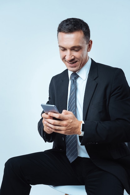 Comunicación online. Hombre agradable encantado alegre sosteniendo su teléfono inteligente y sonriendo mientras envía un mensaje de texto