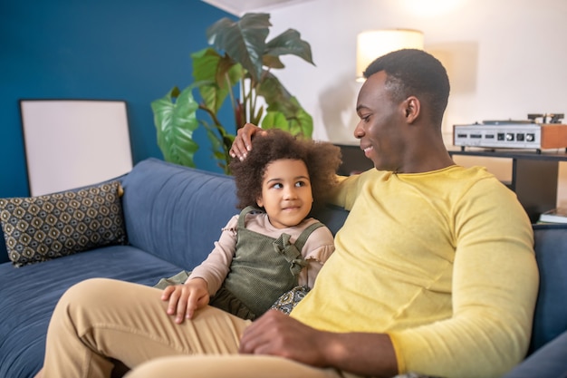 Comunicación. Papá joven de piel oscura tocando la cabeza de la pequeña hija comunicándose sentado en el sofá en la acogedora habitación