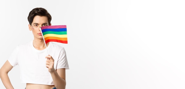 Foto comunidad lgbtq hombre queer atractivo con flitter en la cara ondeando la bandera del arco iris del orgullo y mirando c