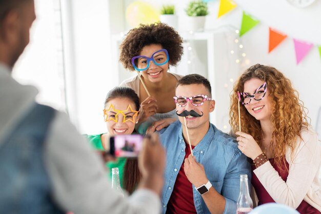 Foto conceito corporativo, de celebração e de feriados - homem tirando uma foto de amigos ou equipe felizes com acessórios de festa se divertindo no escritório