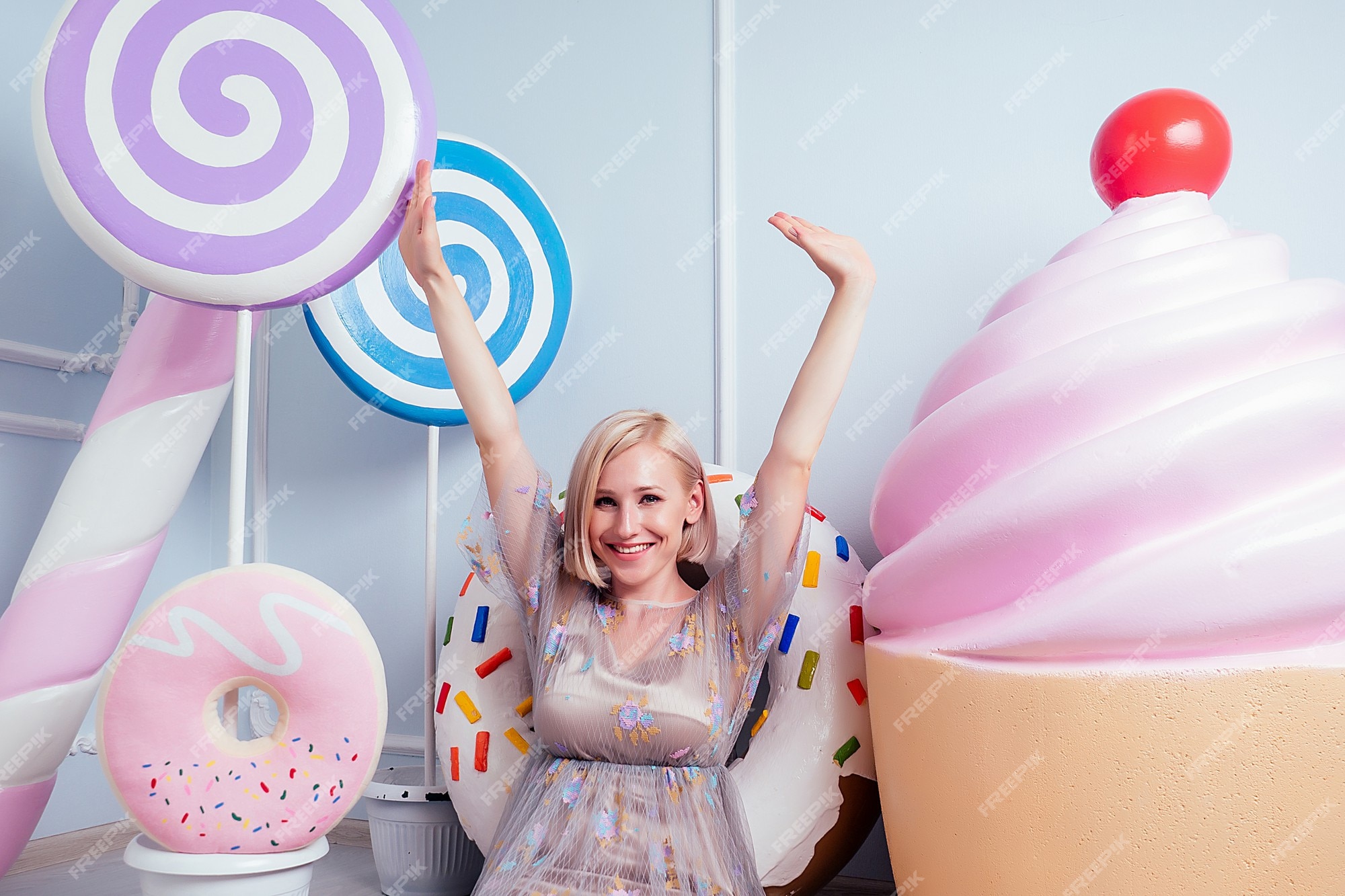 Conceito de adoçamento. linda jovem loira barbie doce mulher confeiteiro modelo  maquiagem perfeita sentada ao lado de pirulito e confeitaria falsa doces  donut doces enormes donuts bolo fundo em estúdio