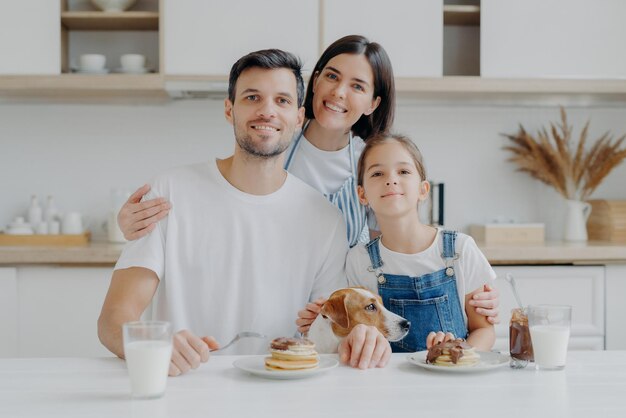 Conceito de alimentação de passatempo de lazer em família Pai mãe e filha jack russell terrier cão posam todos juntos na câmera contra o interior da cozinha têm o prazer de comer panquecas beber leite fresco