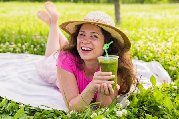 Conceito de alimentação, verão e pessoas saudáveis - uma jovem se diverte no parque e bebe smoothies verdes em um piquenique