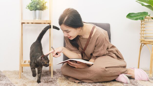 Conceito de amante de animais de estimação Jovem asiática brincando com gato e tomando notas no notebook na sala de estar
