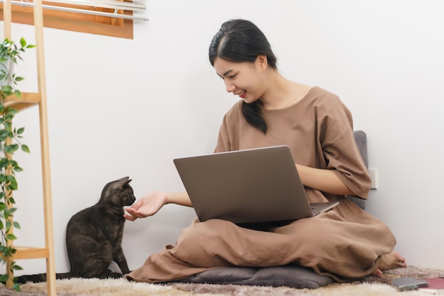 Conceito de amante de animais de estimação Jovem asiática usando laptop e brincando de acariciar com gato na sala de estar