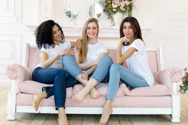 Conceito de amizade e felicidade - três amigas conversando em casa. Três mulheres felizes, engraçadas, multiétnicas melhores amigas rindo e se divertindo, mulheres bem diversas usam camisetas brancas e jeans