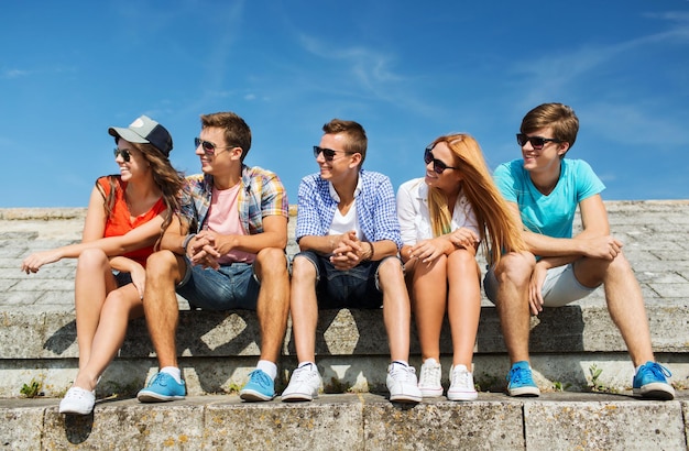 Conceito de amizade, lazer, verão e pessoas - grupo de amigos sorridentes em óculos de sol sentados na rua da cidade