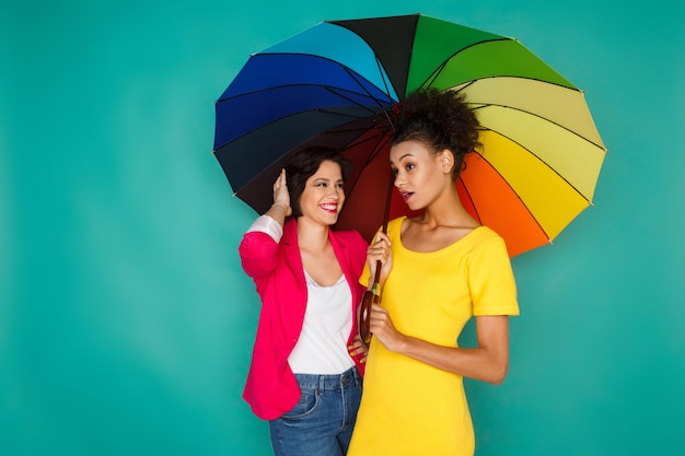 Conceito de amizade multiétnica. Garotas mulatas e brancas com roupas casuais coloridas fofocando sob o guarda-chuva colorido do arco-íris no fundo azul do estúdio com espaço de cópia
