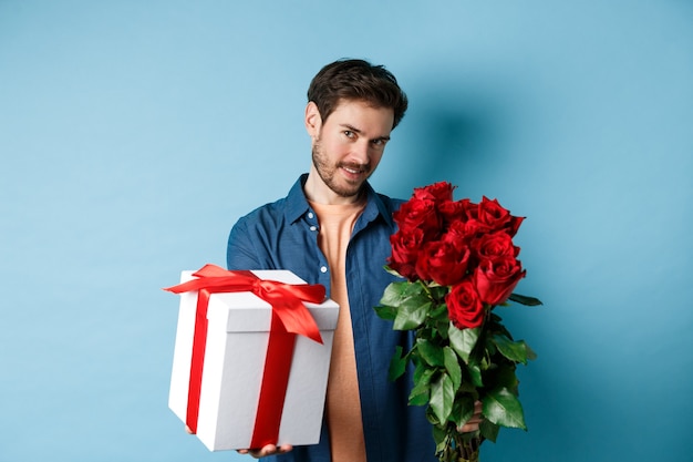 Conceito de amor e dia dos namorados. homem romântico, dando-lhe uma caixa de presente e buquê de flores no encontro, em pé sobre um fundo azul.