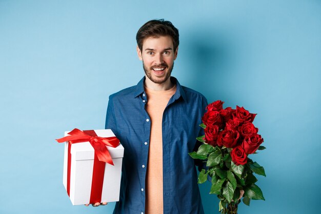 Conceito de amor e relacionamento. Jovem feliz traz flores e presentes em um encontro romântico. Namorado deseja um buquê de rosas e apresentar em pé sobre fundo azul.