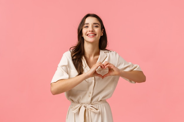 Conceito de amor, ternura e beleza. Moda mulher morena linda no vestido, mostrando sinal de coração, expressar amor e carinho, sorrindo alegre, parede rosa de pé