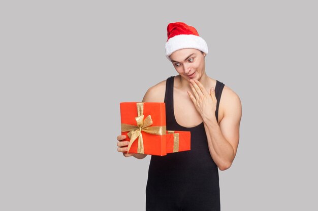 Conceito de ano novo. Homem de chapéu vermelho segurando uma caixa de presente vermelha e olhando para dentro com uma cara engraçada espantada. Interior, foto de estúdio, isolado em fundo cinza