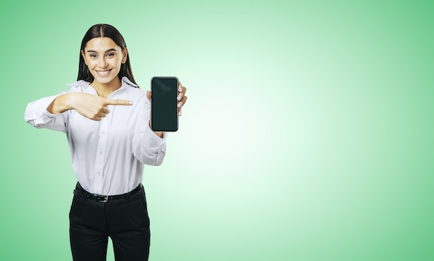 Conceito de aplicativo móvel com garota feliz na camisa branca mostrando celular moderno com tela em branco sobre fundo verde claro abstrato com lugar para o seu logotipo ou maquete de texto
