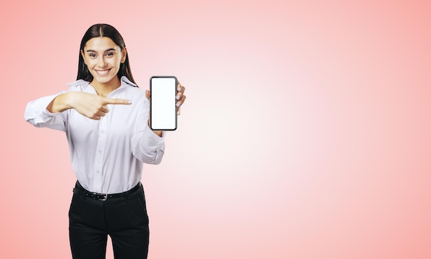 Conceito de aplicativo móvel com garota feliz na camisa branca mostrando smartphone moderno com tela branca em branco de maquete em fundo rosa claro abstrato