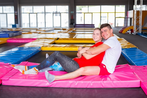 Conceito de atividade física, divertida, de lazer e esporte - casal feliz e alegre sentado em um trampolim dentro de casa