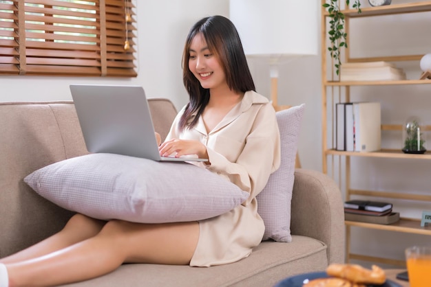 Conceito de atividade na sala de estar Jovem asiática usa laptop para navegar nas mídias sociais no sofá