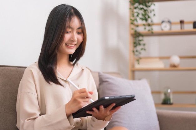 Conceito de atividade na sala de estar Mulher asiática escrevendo em tablet para trabalhar enquanto está sentada no sofá