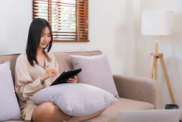 Conceito de atividade na sala de estar mulher asiática escrevendo em tablet para trabalhar enquanto está sentada no sofá