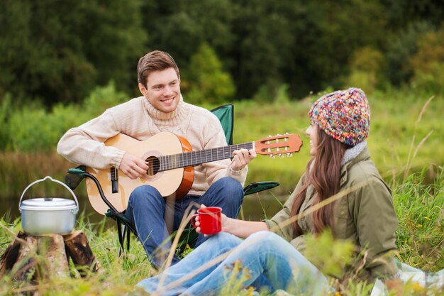 conceito de aventura, viagens, turismo e pessoas - casal sorridente com guitarra cozinhando comida e bebendo no acampamento