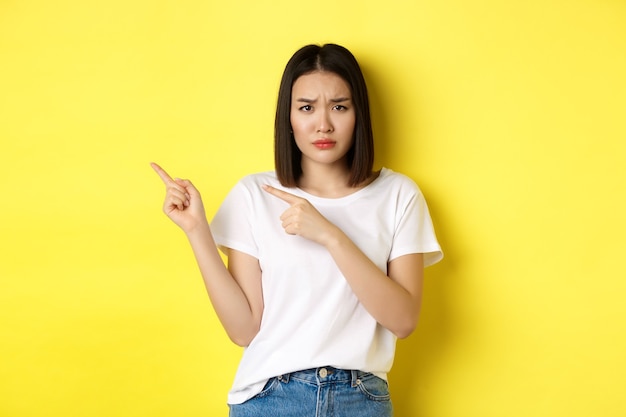 Conceito de beleza e moda. linda mulher asiática em t-shirt branca apontando os dedos para a esquerda, em pé sobre um fundo amarelo.