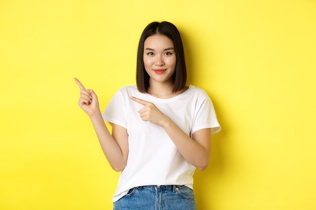 Conceito de beleza e moda. Linda mulher asiática em t-shirt branca apontando os dedos para a esquerda, em pé sobre um fundo amarelo.